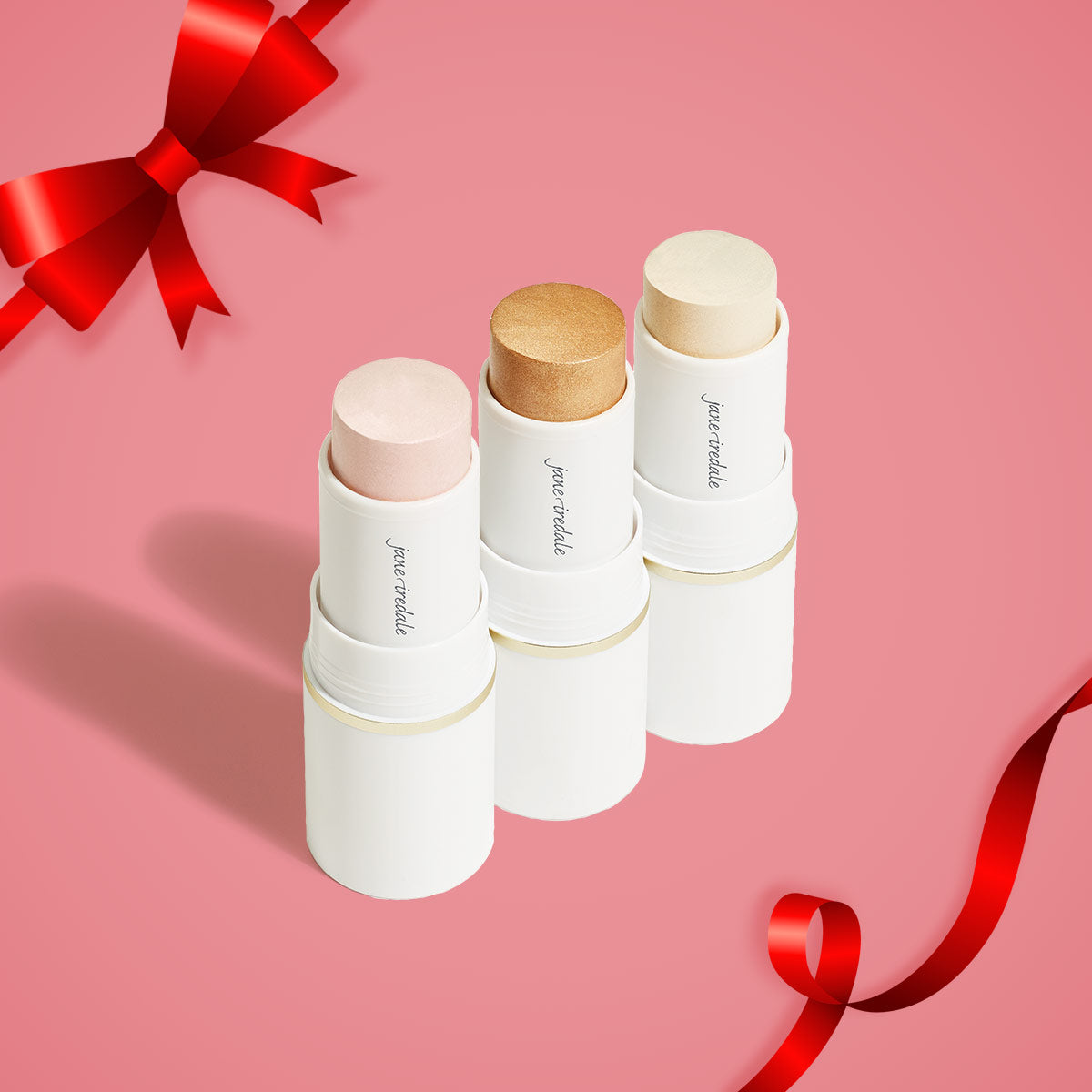 Ein Produktbild des jane iredale Mineral Make-up Produktes Glow Time Highlighter Stick auf einem rosafarbenen Hintergrund und mit einer Schleife in der oberen linken Ecke des Bildes.